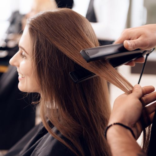 Stylist is Straighten Brown Hair of Woman.